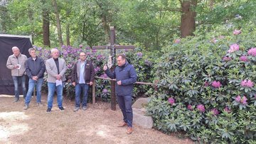Ein Mann steht rechts vor einen Holzkreuz und spricht. Links vom Kreuz stehen weitere vier Männer, sie hören dem Sprecher zu. Im Hintergrund sind blühemde Büsche zu sehen. 
