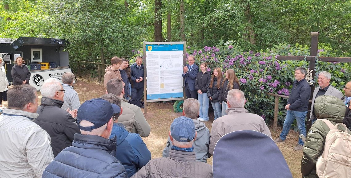 Im Vordergrund eine Gruppe von Menschen, mit dem Rücken zum Fotograf. Dahinter, in der Bildmitte, eine große Tafel mit Abbildungen und Texten. Links und rechts davon einige Menschen verschiedenen Alters.