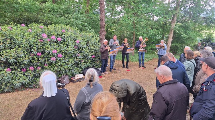 Im Halbkreis mit dem Rücken zum Fotografen eine Gruppe Menschen verschiedenen Alters. Im Hintergrund fünf Personen die Blechinstrumente spielen. Hinter ihnen Büsche und Bäume.