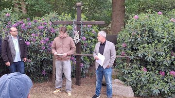 Ein Schüler steht vor einem Holzkreuz. Er trägt von einem Zettel vor. Links und rechts jeweils ein Mann, sie schauen ihn an und hören zu. Rechts im Vordergrund ein Hinterhopf. Im Bildhintergrund Bepflanzung.